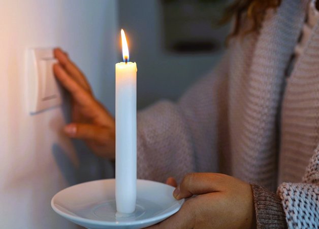 Eine Person mit Kerze in der Hand greift zum Lichtschalter an der Wand. Szene bei Stromabschaltung mit improvisiertem Licht.