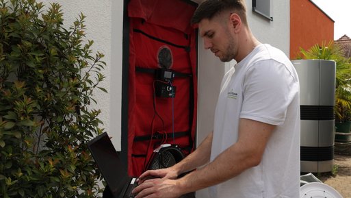 Ein Techniker bedient ein Laptop vor einer Tür mit installierter Blower-Door-Ausrüstung, um Luftdichtheit eines Gebäudes zu testen.