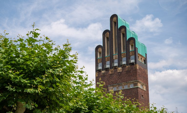 Hochzeitsturm in Darmstadt unter blauem Himmel, umgeben von Bäumen. Bekräftigt Darmstadts Engagement für umweltfreundliche Energie mit Fernwärme.