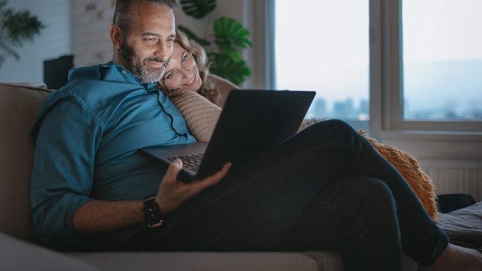 Glückliches Paar sitzt gemütlich auf dem Sofa, schaut auf einen Laptop. Symbolisiert Informationen von ENTEGA zu Rechten von Mietern & Vermietern bei Glasfaser in Mietwohnungen.
