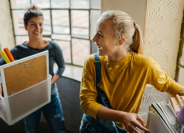 Zwei junge Personen mit Kisten, Pflanzen und Büchern im Treppenhaus. Ihr Ziel: Strom anmelden erste Wohnung.