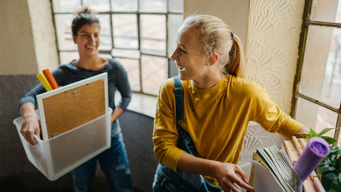 Zwei junge Personen mit Kisten, Pflanzen und Büchern im Treppenhaus. Ihr Ziel: Strom anmelden erste Wohnung.