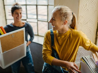 Zwei junge Personen mit Kisten, Pflanzen und Büchern im Treppenhaus. Ihr Ziel: Strom anmelden erste Wohnung.