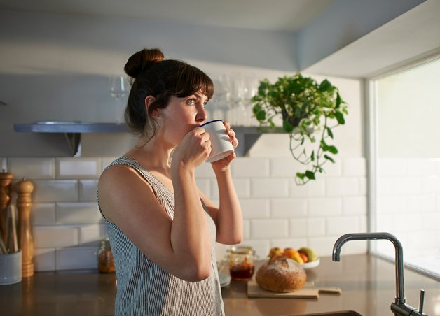 Frau trinkt Tee in heller Küche mit Pflanzen und frischem Brot – klimafreundlich leben durch bewusste Ernährung.