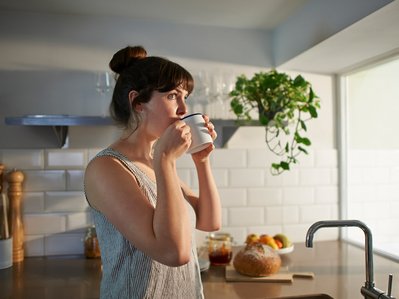 Frau trinkt Tee in heller Küche mit Pflanzen und frischem Brot – klimafreundlich leben durch bewusste Ernährung.