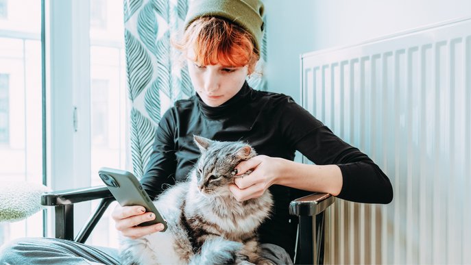 Bild zeigt eine Frau vor einem Heizkörper, auf ihrem Smartphone tippend und mit Katze auf dem Schoß, Symbolbild für Gasheizungswartung 