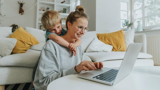 Frau arbeitet am Laptop im Wohnzimmer, Kind umarmt sie von hinten.