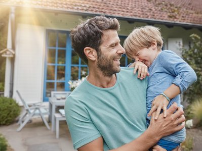 Lachender Vater trägt fröhlichen Jungen im Arm vor einem Haus mit blauem Fensterrahmen. Ausdruck von Freude und familiärer Verbindung an einem sonnigen Tag.