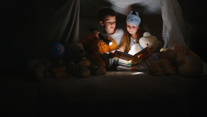 Kinder in Deckenhöhle, umgeben von Kuscheltieren, nutzen Licht von Taschenlampe, symbolisiert Kreativität bei Stromausfall.