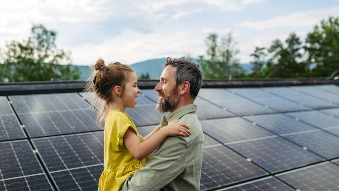 Vater hält Tochter vor Solaranlage im Arm, beide lächelnd. Ausdruck der persönlichen Energiewende und Unabhängigkeit durch Solarenergie.