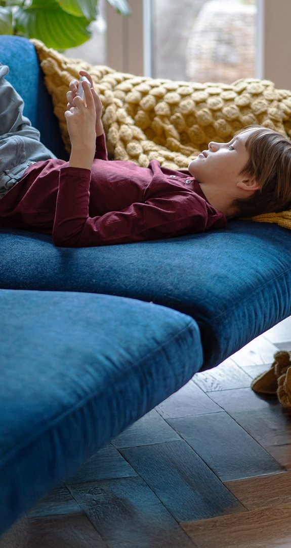 Kind liegt entspannt auf einem blauen Sofa und nutzt ein Smartphone. Eine gelbe Decke ergänzt die gemütliche Atmosphäre im Wohnzimmer.