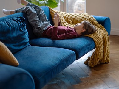 Kind liegt entspannt auf einem blauen Sofa und nutzt ein Smartphone. Eine gelbe Decke ergänzt die gemütliche Atmosphäre im Wohnzimmer.