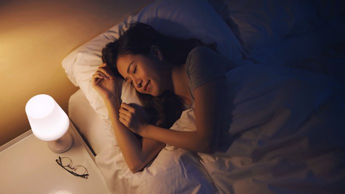 Frau schläft friedlich im Bett neben eingeschalteter Lampe, symbolisiert Vorteile eines Nachtstromtarifs.