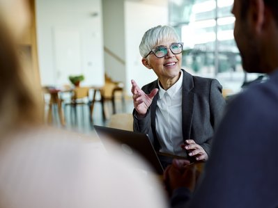 Eine Frau im grauen Blazer diskutiert lebhaft mit Kollegen in einem lichtdurchfluteten Raum, zeigt Dynamik und Professionalität.