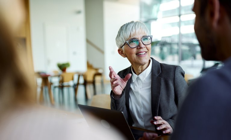 Eine Frau im grauen Blazer diskutiert lebhaft mit Kollegen in einem lichtdurchfluteten Raum, zeigt Dynamik und Professionalität.