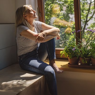 Person genießt Sonnenstrahlen durch das offene Fenster, neben ihr blühende Pflanzen, sommerliche Atmosphäre.