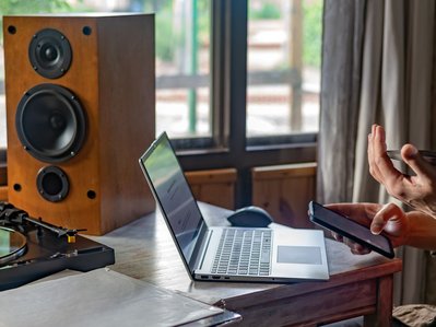 Schreibtisch mit Laptop, Lautsprecher und Plattenspieler. Eine Person informiert sich am Handy zum Thema Stromverbrauch Stand by Geräte.