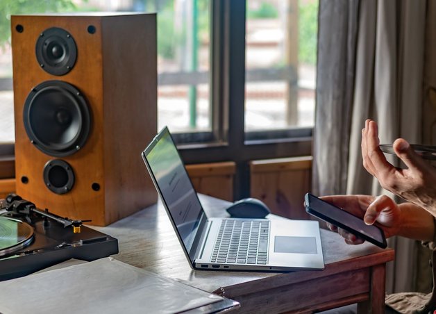 Schreibtisch mit Laptop, Lautsprecher und Plattenspieler. Eine Person informiert sich am Handy zum Thema Stromverbrauch Stand by Geräte.
