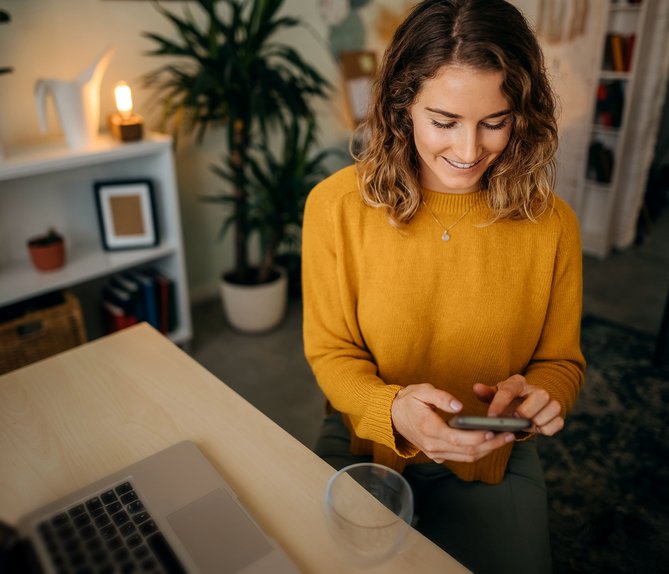 Frau in gelbem Pullover sitzt am Tisch und nutzt ein Smartphone zum Wechsel des Erdgasanbieters. Laptop und Pflanzen im Hintergrund.