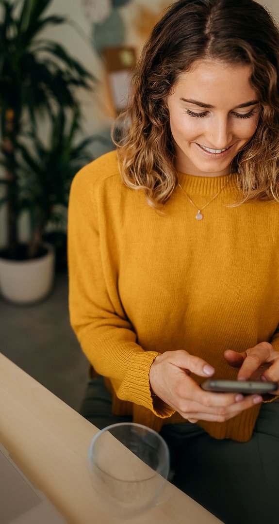 Frau in gelbem Pullover sitzt am Tisch und nutzt ein Smartphone zum Wechsel des Erdgasanbieters. Laptop und Pflanzen im Hintergrund.