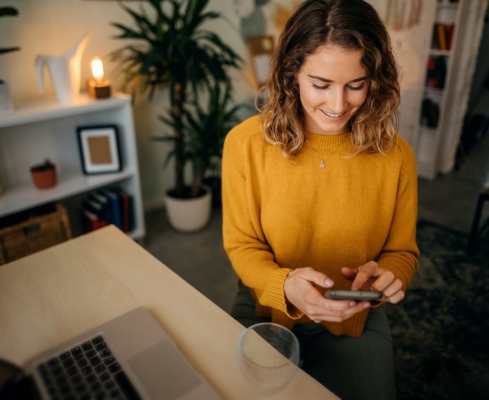 Frau in gelbem Pullover sitzt am Tisch und nutzt ein Smartphone zum Wechsel des Erdgasanbieters. Laptop und Pflanzen im Hintergrund.