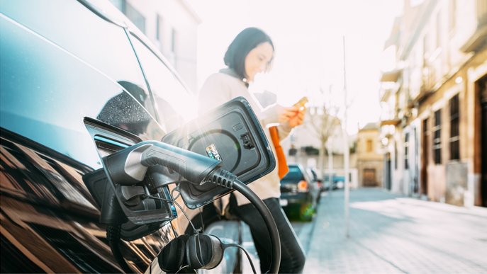 Bild zeigt eine Frau, die ihr Elektroauto auflädt, und dabei etwas auf ihrem Handy tippt