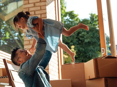 Vater hebt lachende Tochter zwischen Umzugskartons hoch; Umzugskosten werden durch gemeinsames Anpacken und Freude relativiert.