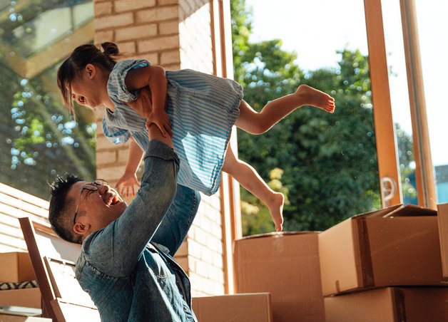 Vater hebt lachende Tochter zwischen Umzugskartons hoch; Umzugskosten werden durch gemeinsames Anpacken und Freude relativiert.