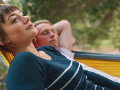 Zwei Personen entspannen in einer Hängematte im Freien. Eine Frau schaut nachdenklich, während ein Mann mit verschränkten Armen ruht. Natürliche Umgebung.