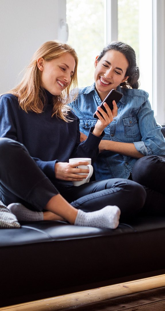 Zwei Personen sitzen entspannt auf einem Sofa, lächeln und schauen auf ein Smartphone in einer gemütlichen Wohnraumumgebung.