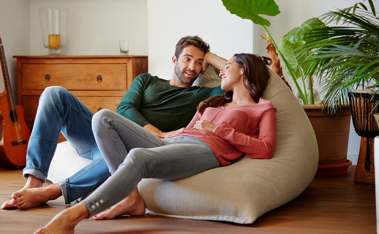 Zwei Personen entspannen sich auf einem Sitzsack in einem gemütlichen Wohnzimmer mit Pflanzen und einer Gitarre im Hintergrund.